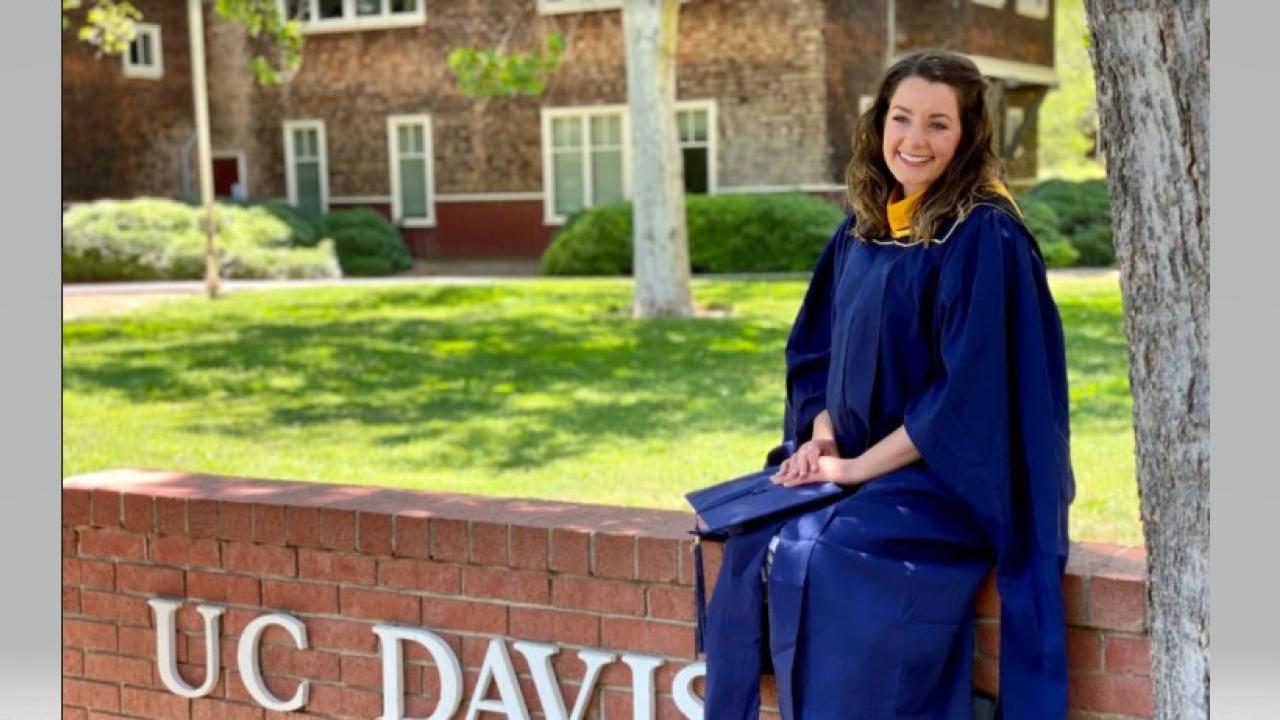 Picture of Amanda in her graduation gown sitting on a brick wall that says UC DAvis