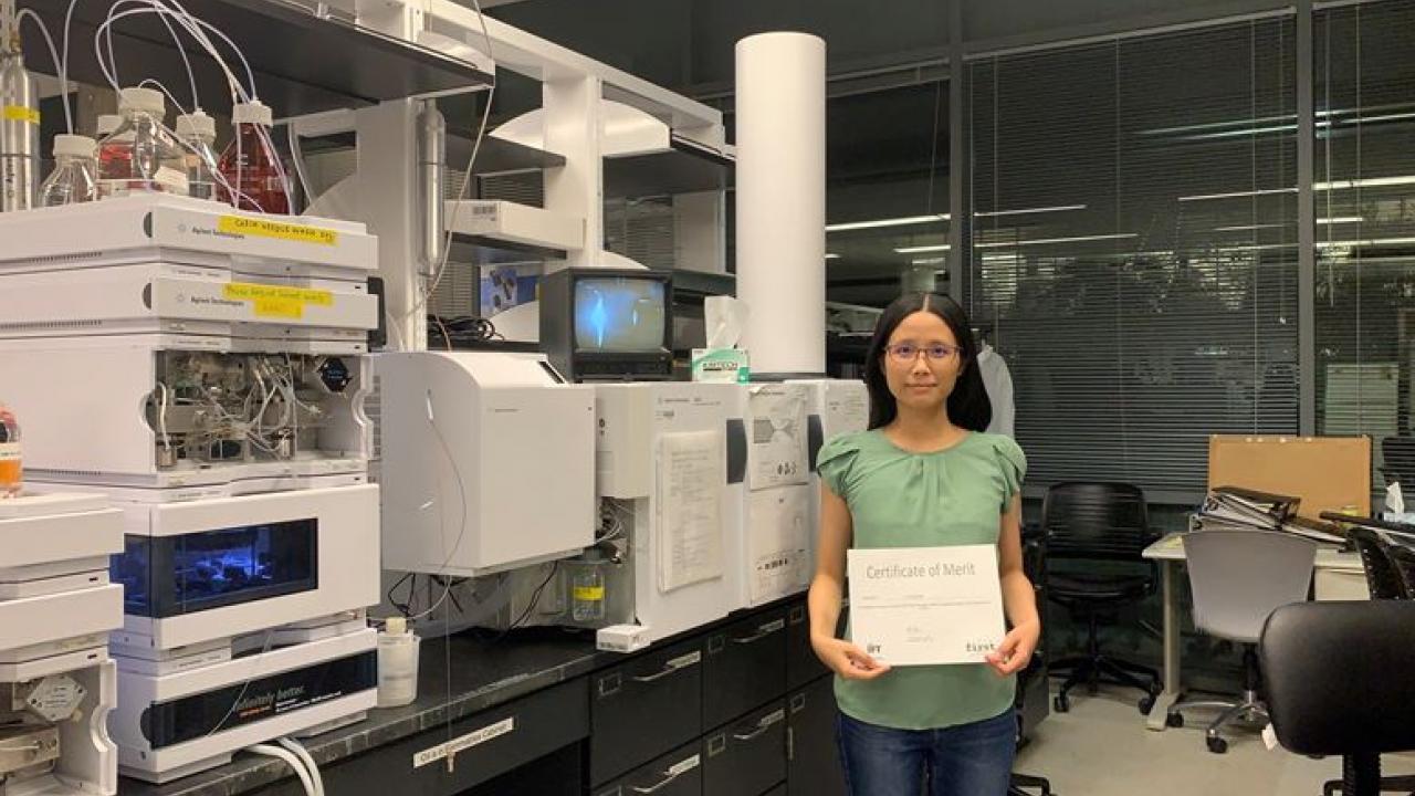 Picture of Yu-Ping holding a certificate standing in front of the LC-Q-ToF in the Barile Lab